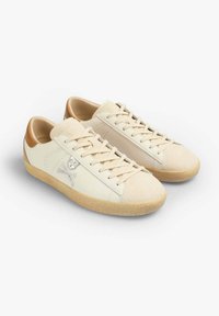 Cream-colored sneakers with a smooth leather body, suede toe cap, and tan accents. Features lace-up closure and a skull and crossbones logo.