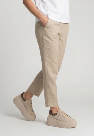 Person wearing beige cropped pants and beige platform sneakers, standing with one hand in pocket against plain background.