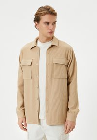 Chemise beige texturée avec un col, fermeture à boutons sur le devant et deux poches poitrine. Présente des manches longues avec des poignets à boutons.