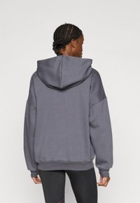 Pilkas džemperis su laisvu fitu, su raišteliu reguliuojama gaubtuvu, nuleistomis pečių siūlėmis ir ribinėmis rankogaliais apačioje ir rankovėse.