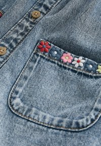 Poche de veste en denim avec des fleurs brodées colorées le long du bord supérieur et des boutons en bois sur la patte de boutonnage.
