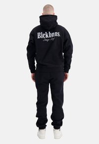 Zwarte hoodie en joggingbroek set van katoen, met een groot geborduurd logo op de achterkant. Witte schoenen met een geprofileerde zool.