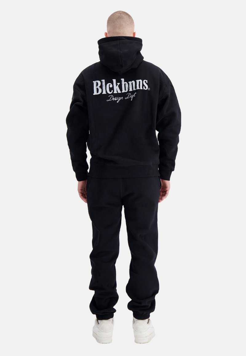 Zwarte hoodie en joggingbroek set van katoen, met een groot geborduurd logo op de achterkant. Witte schoenen met een geprofileerde zool.