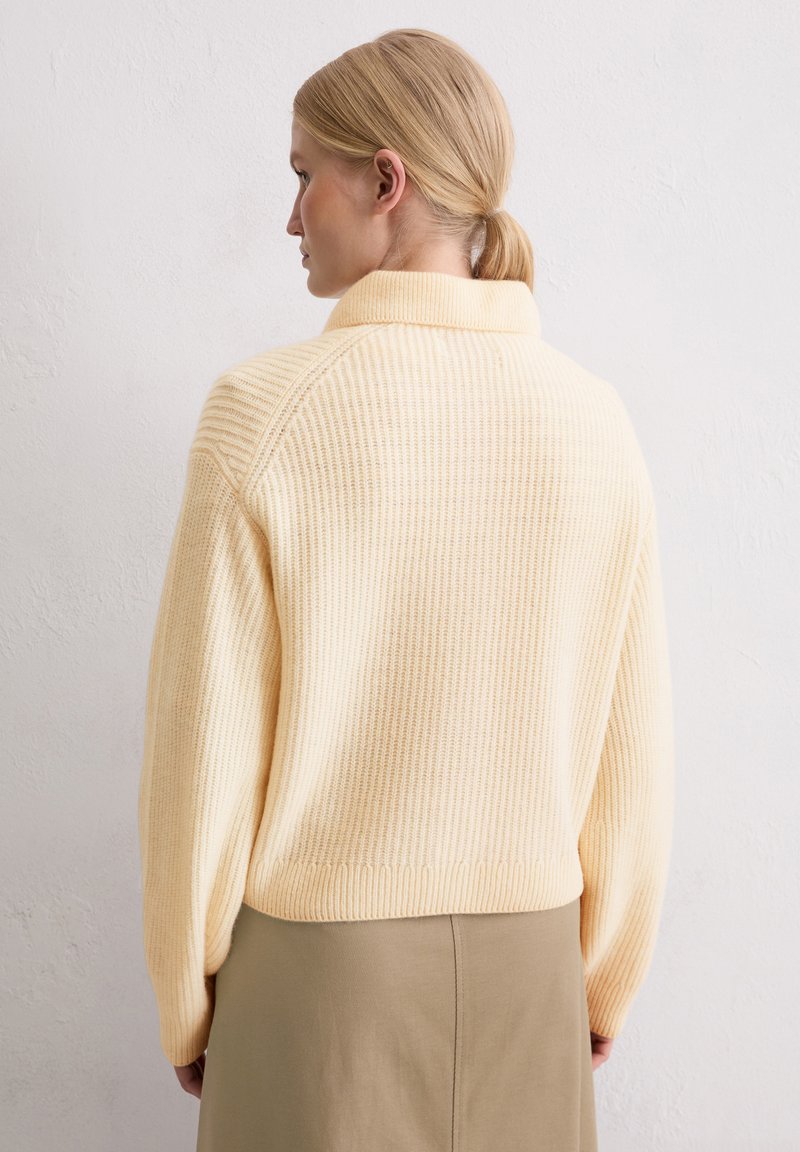 Hellgelber, gerippter Pullover mit hohem Kragen, lockerem Schnitt und überschnittenen Schultern, kombiniert mit einem schlichten Rock. Weiche Textur, minimalistische Details.