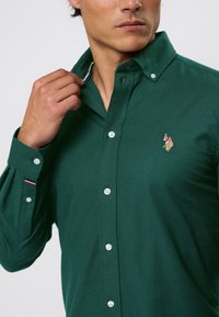 Zöld pamut gombos ing, tűzött gallérral, csíkozott díszítéssel és egy kis hímzett logóval a mellrészen.