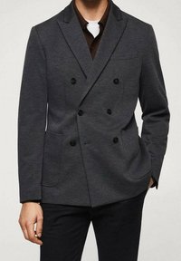 Blazer grigio doppiopetto con bottoni neri, revers a punta, due tasche frontali e un tessuto elasticizzato. Indossato sopra una camicia scura.