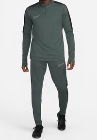 Donkergroene sportieve trainingspak met een ritskraag, lange mouwen en zwarte accenten aan de zijkanten, gemaakt van lichtgewicht, gestructureerde stof. Nike-logo aanwezig.