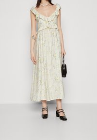 Robe maxi à imprimé floral en crème clair avec des manches volantes et une taille froncée. Associée à des escarpins noirs à bout fermé et à un petit sac noir.