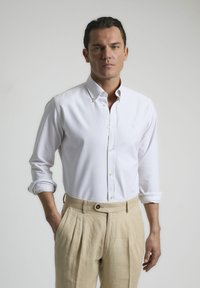 Camisa blanca de botones con rayas verticales claras, puños arremangados y cuello, combinada con pantalones beiges con pliegues y detalle de botón.