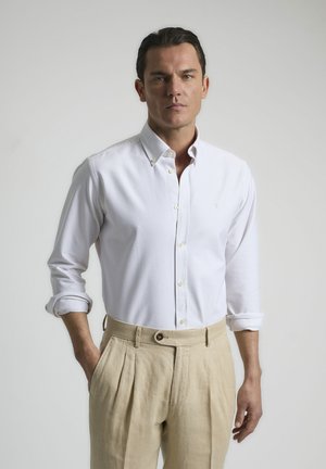 Camisa blanca de botones con rayas verticales claras, puños arremangados y cuello, combinada con pantalones beiges con pliegues y detalle de botón.