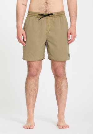 Homme portant un short de bain beige mi-cuisses avec un cordon noir, debout pieds nus devant un fond blanc uni.