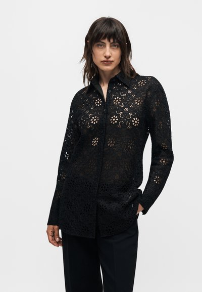 Γυναίκα με σκούρα μαλλιά που φοράει μαύρο πουκάμισο από δαντέλα με λουλούδια και μαύρο παντελόνι, στέκεται με το ένα χέρι στην τσέπη μπροστά σε λευκό φόντο.