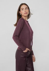 Cárdigan marrón hecho de una tela suave, con mangas largas, cuello redondo y cierre de botones. Combinado con pantalones de cuero oscuros.