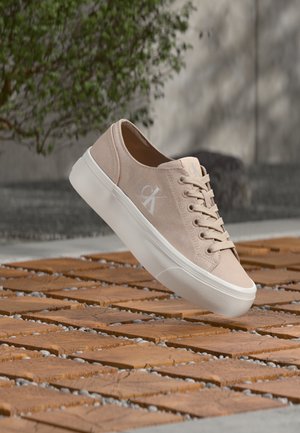 Jasnobeżowy tenisówka z płótna z białą gumową podeszwą, wyposażona w sznurowadła i małe logo po boku. Teksturowana powierzchnia i minimalistyczny design.
