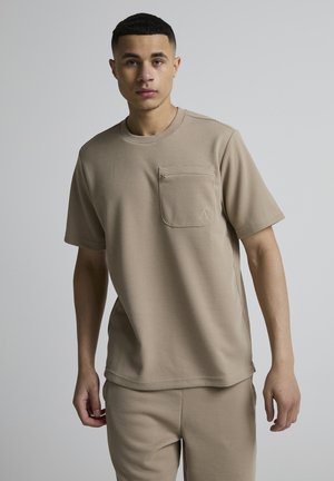 Jonge man staand tegen een effen grijze achtergrond, gekleed in een beige, getextureerd shirt met korte mouwen met een borstzak en bijpassende broek.