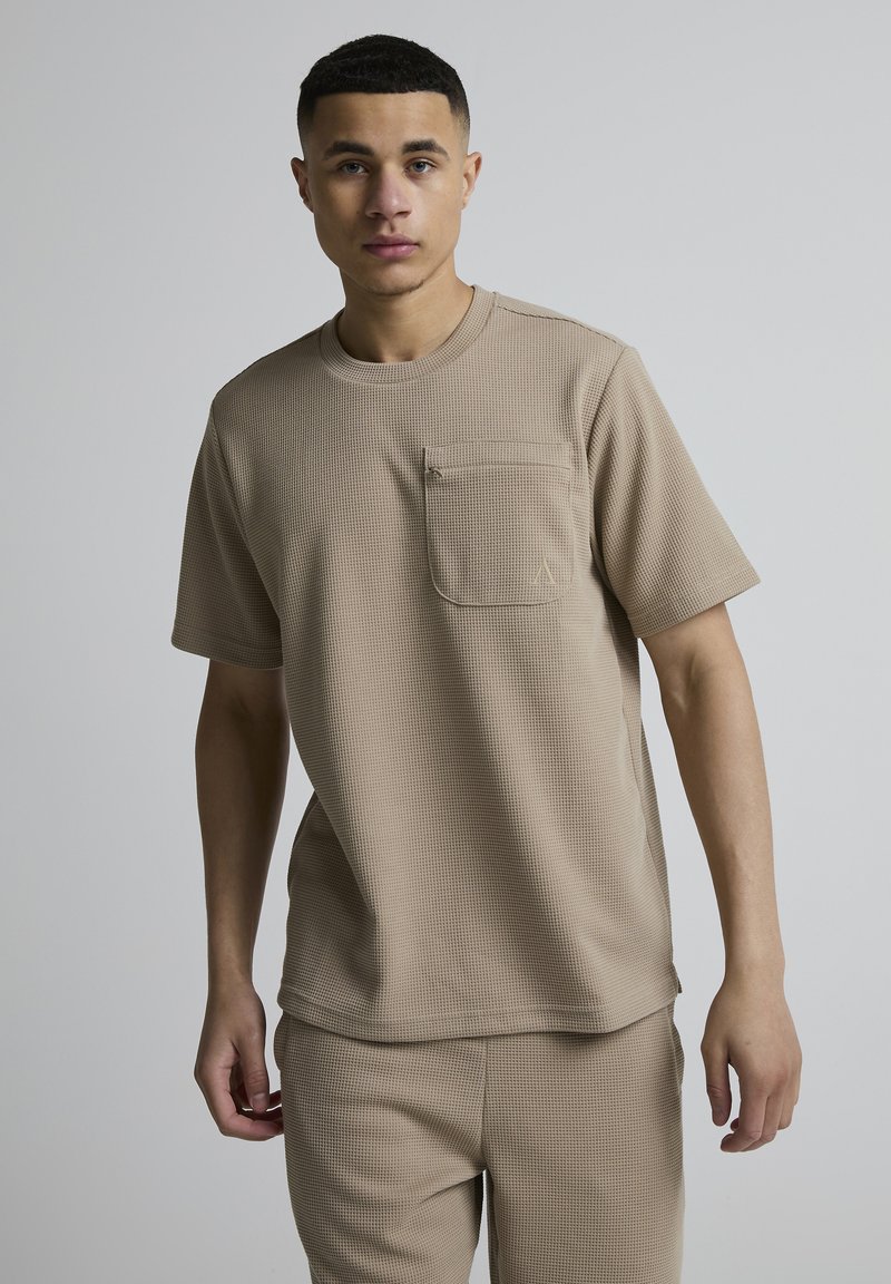 Jeune homme debout devant un fond gris uni, portant une chemise beige texturée à manches courtes avec une poche poitrine et un pantalon assorti.