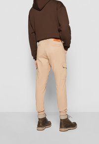Pantalon cargo beige avec poignets élastiques, deux poches latérales et une écusson à l'arrière. Associé à un sweat à capuche marron foncé et des bottes.