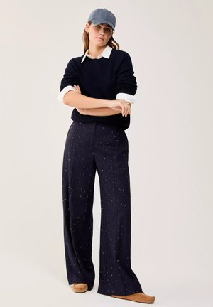Donna in piedi con le braccia incrociate, indossa un maglione blu navy sopra una camicia bianca con colletto, pantaloni larghi blu navy a pois, berretto blu e mocassini marroni.