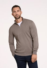 Brauner Sweatpullover mit Reißverschluss und glattem Strickstoff, mit langen Ärmeln und geripptem Saum, kombiniert mit marineblauen Hosen vor einem neutralen Hintergrund.