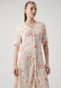 Rochie cu flori, cu maneci scurte, de tip cămașă, având un decolteu în V și un fundal roz pal decorat cu modele florale colorate.