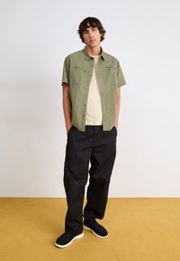 Chemise à manches courtes, vert clair, avec deux poches poitrine, superposée sur un t-shirt beige, assortie à un pantalon noir ample et des chaussures noires texturées.