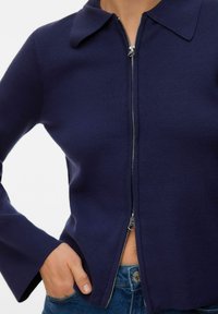 Cardigan bleu marine à fermeture éclair, avec col, texture côtelée, manches longues, et détails en fermeture éclair argentée, porté sur un jean bleu.