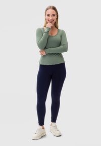 Camicia verde a maniche lunghe con bordo in pizzo e leggings blu navy. L'outfit è abbinato a sneakers bianche su uno sfondo semplice.