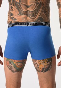Blå bomullsboxershorts med svart midja som har "JP1880"-logotyp. Synliga tatueringar på låren inkluderar detaljerade drake- och tigerdesigns.