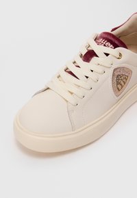 Baskets créme avec de grosses lacets blancs, une languette en velours bordeaux et un patch beige orné de strass sur le côté.