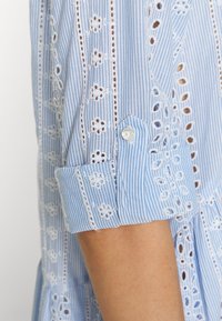 Camisa riscada em azul claro com bordado em fio branco. Possui mangas arregaçadas e um botão de pérola no punho. Textura de algodão suave.