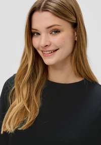 Zwarte sweatshirt met een ronde hals en textuurnaadwerk. Model heeft lang, golvend blond haar en een neutrale achtergrond.