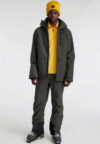 Tamsiai žalia nepralaidi vandeniui striukė su gobtuvu, užtrauktuku priekyje ir šoninėmis kišenėmis, derinama su ryškiai geltonu marškinėliu ir atitinkančia kepure. Juodi akcentai ant kelnių.