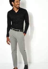 Zwarte long-sleeve shirt met knopen, gecombineerd met lichtgrijze broek en een zwarte riem. De man staat casual, met zwarte schoenen aan.