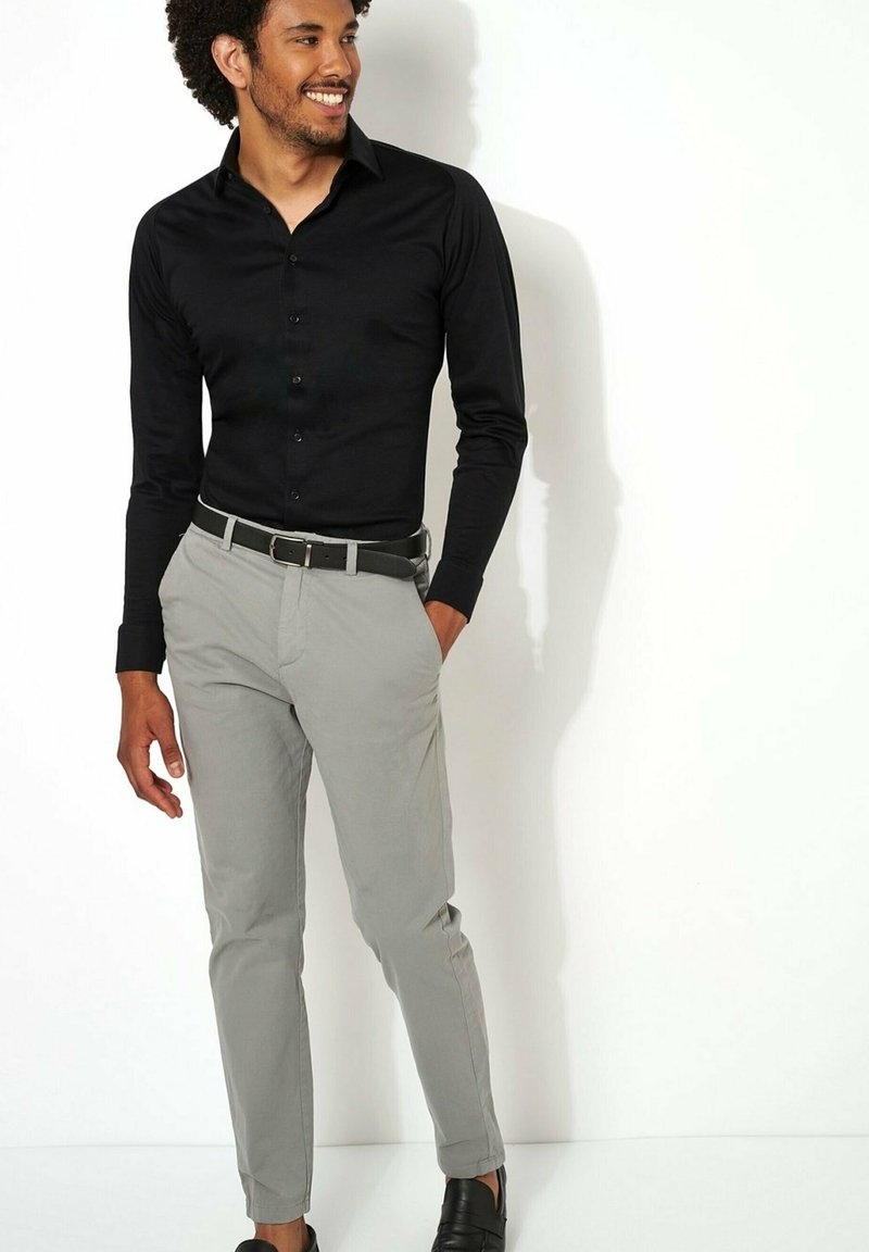 Zwarte long-sleeve shirt met knopen, gecombineerd met lichtgrijze broek en een zwarte riem. De man staat casual, met zwarte schoenen aan.