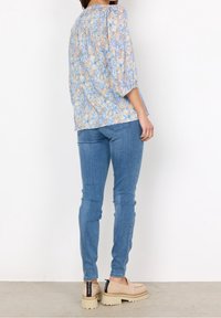 Blusa floreale azzurro chiaro con maniche a tre quarti, abbinata a jeans blu attillati e scarpe con piattaforma beige con un'etichetta nera sul tallone.
