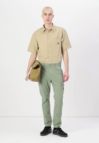 Chemise beige à manches courtes avec une poche poitrine, assortie à un pantalon cargo vert clair avec des poches latérales, et des baskets noires accompagnées d'un sac beige.