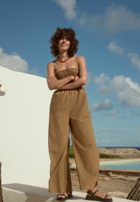 Brun, ärmlös jumpsuit med en smockad bodice och vida ben. Har ett texturerat tyg och en avslappnad passform. Bärs med svarta sandaler.