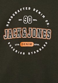Tela de un verde oscuro con texto en blanco y naranja. El logotipo central incluye "JACK & JONES", "HANDCRAFTED DENIM CO." y "EST. 90."