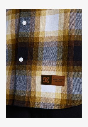 Yellow, gray, and white plaid flannel shirt with white buttons and a brown leather label detailing "QUALITY GOODS SINCE 1994."