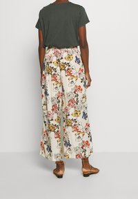 Pantalons larges fleuris en crème, ornés de motifs de fleurs multicolores. Associés à un haut à manches courtes vert foncé et des sandales marron.