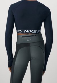 Takaa näky henkilöstä, joka on pukeutunut mustaan, pitkähihaiseen Nike Pro -paitaan ja korkeavyötäröisiin tummanharmaisiin Nike-leggingseihin, joissa on vetoketjullinen tasku.