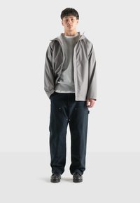 Giacca impermeabile grigia sopra un felpa grigia, abbinata a pantaloni larghi blu scuro. Indossata con scarpe nere, presentata su uno sfondo neutro.