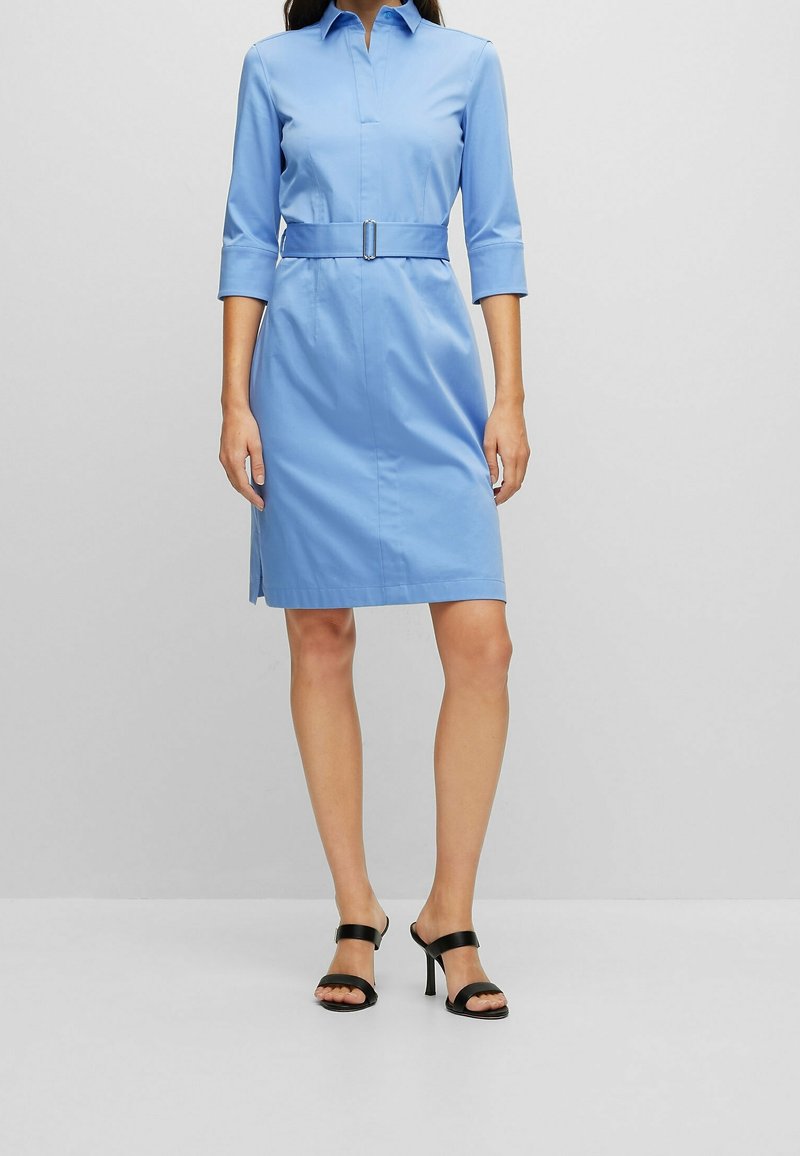Femme portant une robe ceinturée bleu clair longueur genou avec des manches trois-quarts et des sandales noires à talons ouverts.