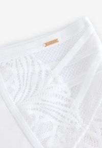 Top blanco con un panel de encaje con un patrón floral texturizado y finas tiras blancas. Cerca del escote se puede ver una pequeña etiqueta metálica de la marca.