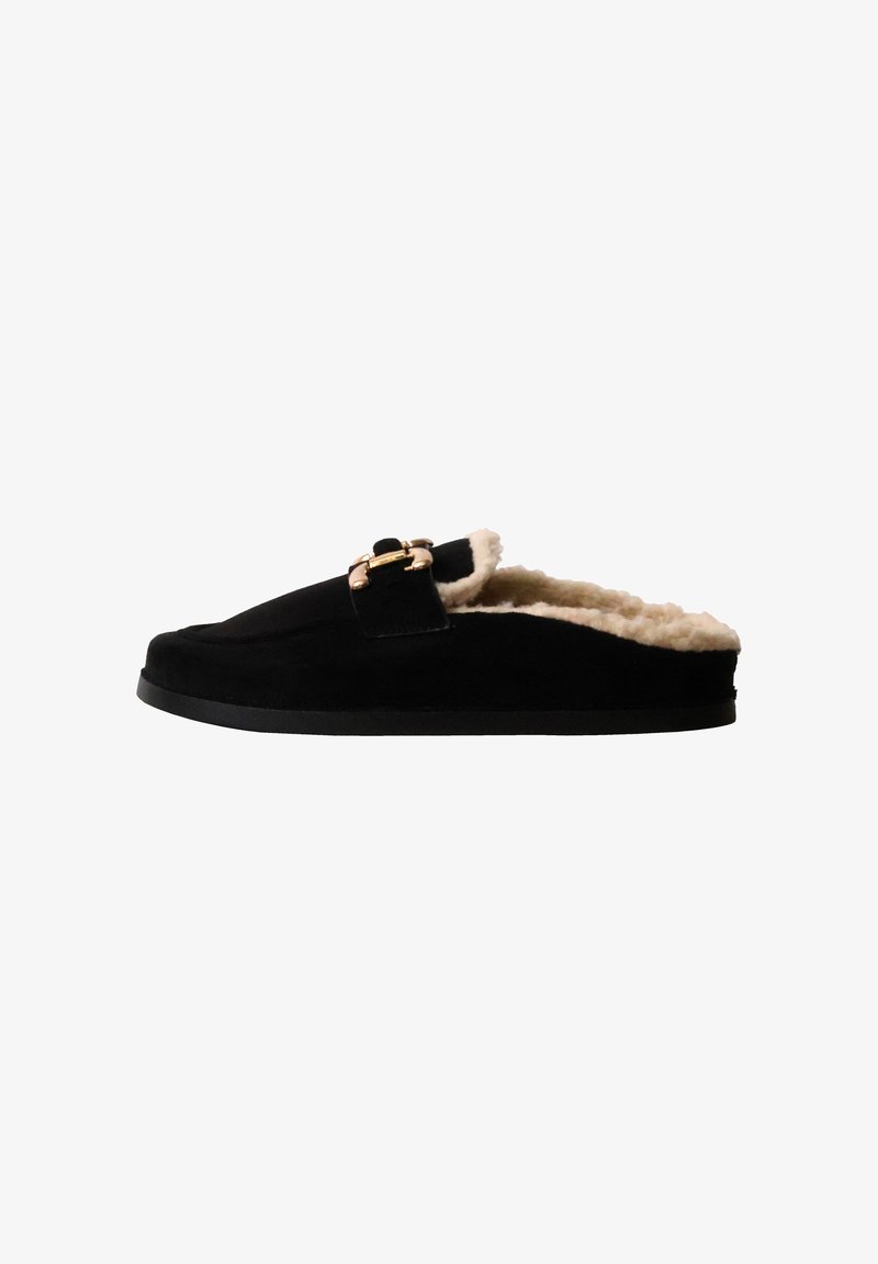 Chausson noir à enfiler avec doublure en fourrure beige et détail de boucle dorée sur le dessus, semelle plate, vue latérale sur fond blanc.