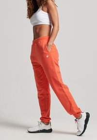 Orange sweatpants med en avslappnad passform, elastisk midja och muddar; kombinerat med vita sneakers med färgglada detaljer.