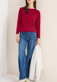 Kobieta w długim, czerwonym topie i niebieskich dżinsach, trzymająca jasnoszary sweatshirt, stojąca przy jasnobeżowej ścianie.
