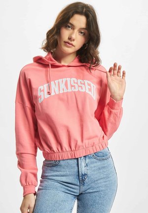 Rosa verkürzter Kapuzenpullover mit weißem "SUNKISSED"-Text, Kordelzug-Kapuze, geripptem Saum und lockerer Passform. Getragen mit hellblauen Jeans.