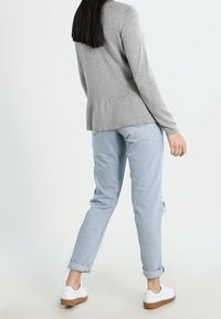 Graues Langarmshirt mit plissiertem Rücken, kombiniert mit hellblauen hochgekrempelten Jeans und weißen Schuhen mit brauner Sohle.