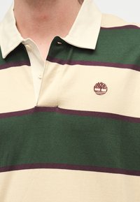 Άτομο που φοράει πουκάμισο με γιακά, με φαρδιές οριζόντιες πράσινες, κρεμ και μωβ ρίγες και ένα μικρό κεντημένο λογότυπο δέντρου στο στήθος.